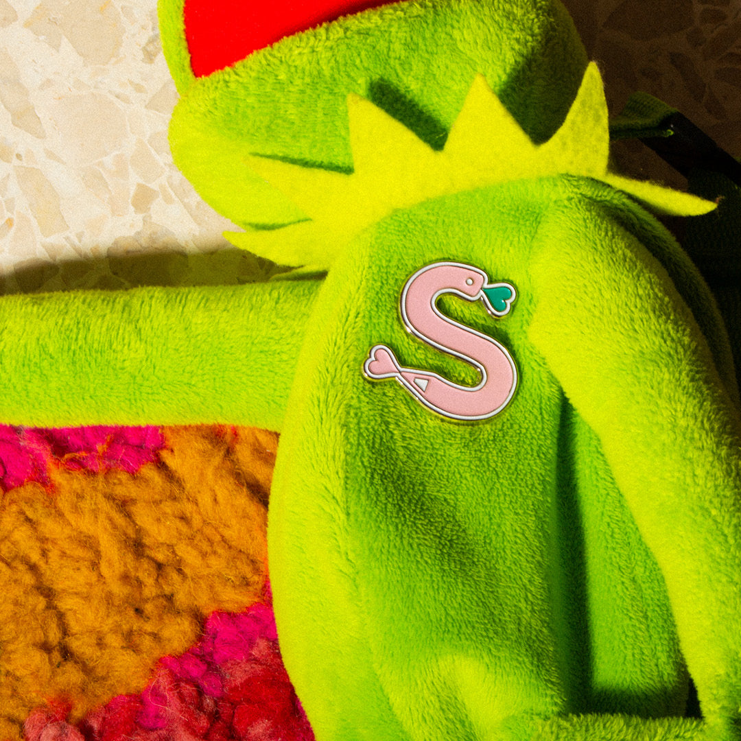 A pink enamel pin shaped like a curled Scribble snake attached to a Kermit the Frog bag, resting on a colorful woven rug.