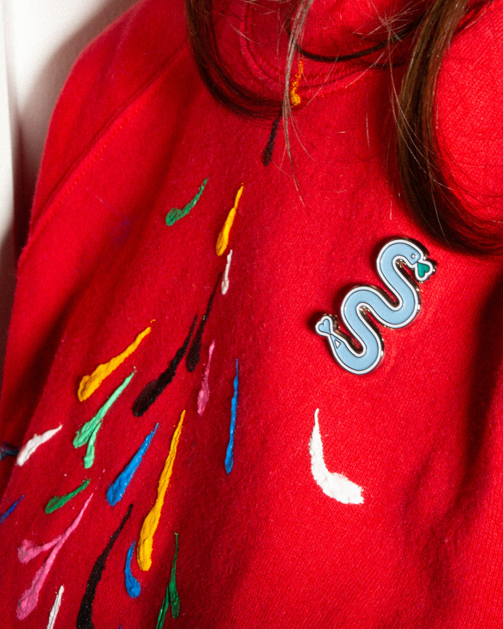 A person wearing a bright red sweatshirt decorated with colorful markings and wearing a wedgwood blue double scribble enamel snake pin