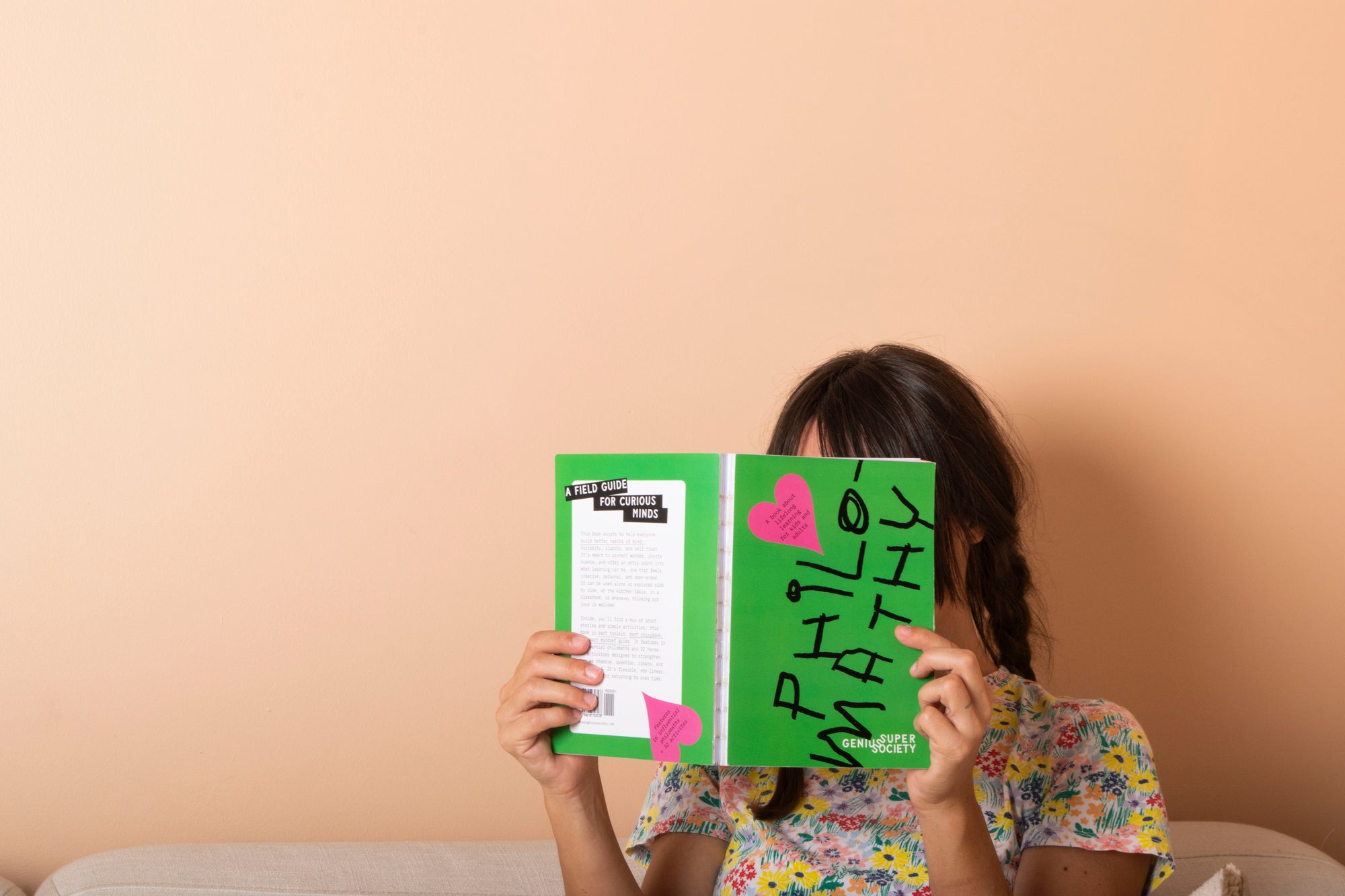 Person reading the Philomathy book with a vibrant green cover and bold graphic black text and a pink heart heart with a view of the back cover that says "A field guide for curious minds" against a pink-beige background