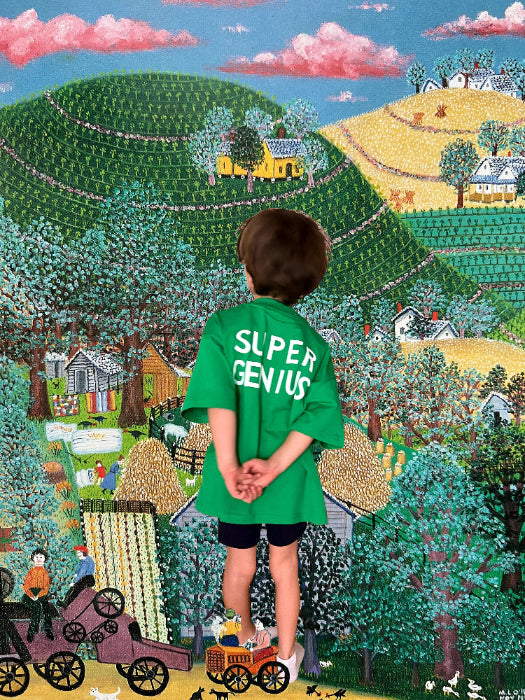 Child wearing a 'Super Genius' shirt standing in front of a colorful, detailed painted landscape.